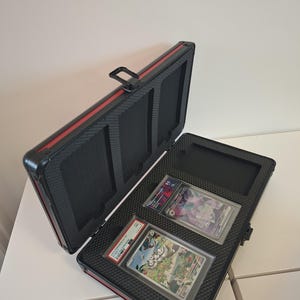 Puede incluir: Malet&iacute;n negro y rojo para tarjetas, abierto para revelar dos cartas coleccionables calificadas. El malet&iacute;n tiene un interior texturizado y un asa. Las tarjetas est&aacute;n en fundas protectoras.