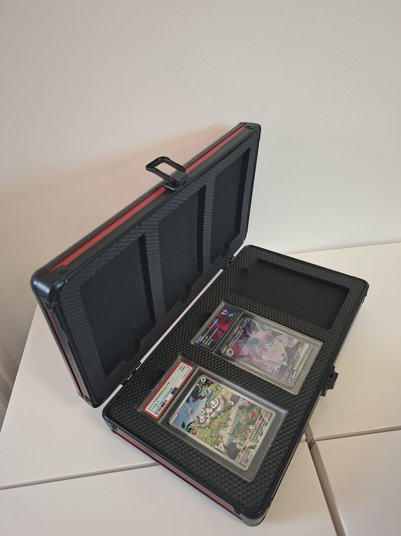 Puede incluir: Malet&iacute;n negro y rojo para tarjetas, abierto para revelar dos cartas coleccionables calificadas. El malet&iacute;n tiene un interior texturizado y un asa. Las tarjetas est&aacute;n en fundas protectoras.