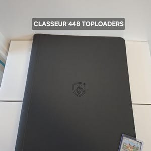 May include: A dark gray binder with the text "CLASSEUR 448 TOPLOADERS" at the top. The binder has a logo in the center and a trading card in a protective sleeve is visible. The binder is designed to hold trading cards.