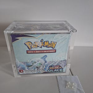 Può includere: Una vetrina trasparente in acrilico che contiene una scatola di carte collezionabili Pokémon. La scatola presenta il logo Pokémon e il testo "Cartes à Jouer et à Collectionner" e "Règne de Glace". Un panno bianco e un piccolo pezzo di plastica sono davanti.