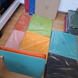 May include: A collection of square, zipped portfolios in various solid colours including red, blue, beige, orange, lime green, pink, yellow, turquoise, and black. Each portfolio features a centred shield logo with a horse emblem on the front cover.