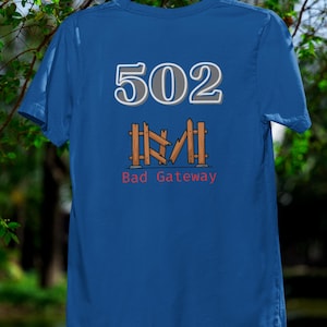 May include: A blue t-shirt with the number "502" above a fence graphic and the words "Bad Gateway" in red. The shirt is hanging on a black hanger against a blurred green background.