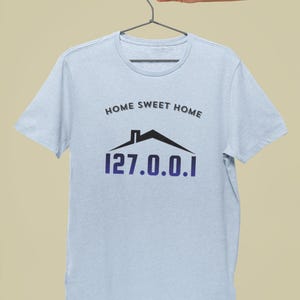 Peut inclure: T-shirt bleu clair avec l'inscription "HOME SWEET HOME" au-dessus d'un dessin de maison noire et l'adresse IP "127.0.0.1" en violet. Le t-shirt est suspendu à un cintre en métal.