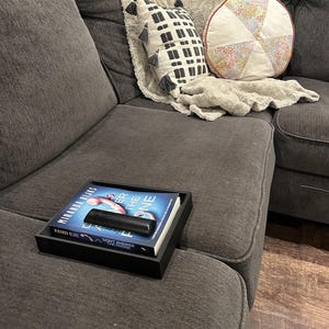 May include: A black rectangular tray holding a book titled Don't Answer the Phone by Miranda Rijks and a black remote control. The tray is placed on a dark gray textured sofa with a white patterned throw blanket and two decorative pillows.
