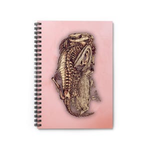 Puede incluir: Cuaderno de espiral con una ilustración detallada de un esqueleto de dinosaurio sobre un fondo rosa. La estructura esquelética está representada en tonos marrones y beige, con detalles intrincados. La encuadernación en espiral del cuaderno es negra.