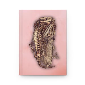 Puede incluir: Un diario rosa con una ilustración detallada de un esqueleto de dinosaurio abrazando a una pequeña criatura peluda. La obra de arte está en tonos marrones y beige, creando una estética vintage. La portada del diario es de color rosa liso.