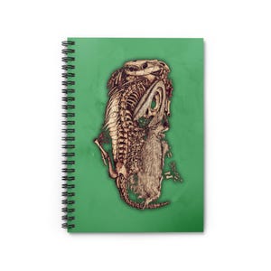 Puede incluir: Cuaderno de espiral con una cubierta verde. La cubierta presenta una ilustración detallada de un esqueleto de dinosaurio y una criatura peluda, en tonos marrones y beige. El cuaderno tiene una encuadernación en espiral negra.