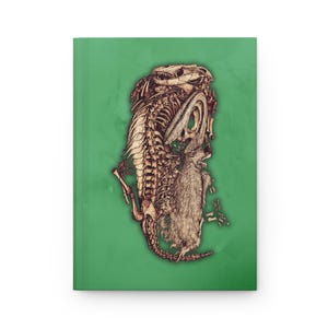 Puede incluir: Un diario de tapa dura con una cubierta verde. El diario presenta una ilustración detallada de un reptil esquelético y un conejo, representados en tonos marrones y beige. La obra de arte está centrada en la portada.