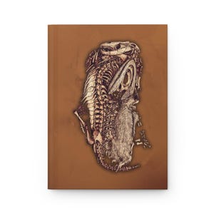 Puede incluir: Un diario marrón con una ilustración detallada de esqueletos de animales, incluyendo un cráneo de reptil y una criatura peluda. La portada es de color marrón liso, y la obra de arte está en tonos marrones y beige.