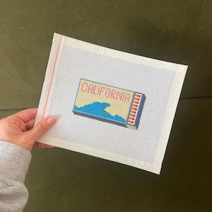 May include: A needlepoint canvas featuring the word "CALIFORNIA" above a blue wave design. The canvas is white with a grid of small holes for stitching. The design includes yellow, blue, and red colors.