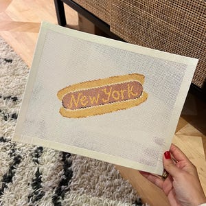 May include: A needlepoint canvas featuring a hot dog design with the text "New York" in a retro style. The hot dog is yellow and brown, set against a cream-colored background. The canvas is held by a person.