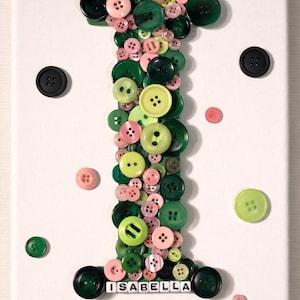 May include: A canvas art piece featuring the letter "I" crafted from various sizes of green and pink buttons. The name "ISABELLA" is spelled out at the bottom. Additional buttons are scattered around the white canvas.