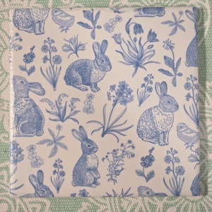 May include: Square ceramic tile with a repeating pattern of blue rabbits, flowers, and foliage on a white background. The design is reminiscent of a vintage botanical illustration. The tile is likely used for decorative purposes.
