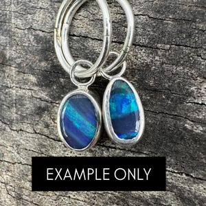 Può includere: Orecchini a cerchio in argento con pendenti in opale di forma ovale e a goccia. Gli opali mostrano vibranti sfumature blu e verdi. Il testo "EXAMPLE ONLY" è su un rettangolo nero.