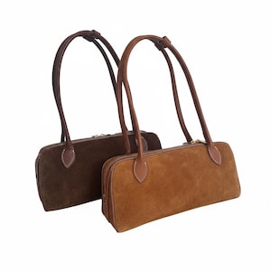 May include: Two suede shoulder bags in shades of brown. The bags have long, curved handles and a rectangular shape with a zipper closure. The stitching is visible, and the handles are a darker brown than the bag bodies.