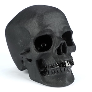 Puede incluir: Una escultura de cráneo negro mate con una superficie texturizada, cuencas oculares profundas y huecas, una cavidad nasal y un conjunto de dientes negros lisos y brillantes. El objeto presenta un acabado oscuro y sólido con contornos anatómicos definidos.