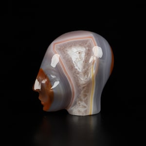 Puede incluir: Una escultura de cabeza tallada en ágata con un patrón de bandas blancas, grises y naranjas. La cabeza está de perfil y tiene un perfil detallado.