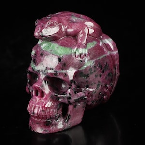 May include: A pink and green gemstone skull with a carved frog on top. The skull is made of ruby zoisite, a type of gemstone known for its vibrant colors and unique patterns.