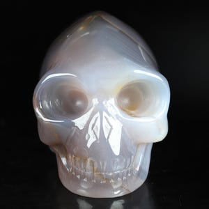 Puede incluir: Una figura de calavera tallada en piedra de ágata translúcida, de color gris claro y blanco. La calavera tiene detalles, incluyendo cuencas de los ojos, una cavidad nasal y dientes. La superficie pulida refleja la luz, creando reflejos y sombras.