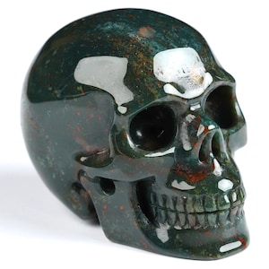 Puede incluir: Una escultura de calavera pulida tallada en piedra verde y moteada de rojo. La calavera tiene rasgos detallados, incluyendo cuencas de los ojos, una cavidad nasal y una dentadura completa. La piedra tiene un acabado brillante, sobre fondo blanco.