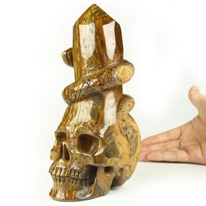 Op de afbeelding: Een gebeeldhouwde stenen sculptuur met een schedel met een slang gewikkeld rond een kristallen punt. De sculptuur is voornamelijk bruin en goud, met gedetailleerde gravures. De schedel heeft zichtbare tanden en oogkassen.