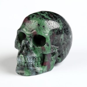 以下が含まれることがあります： 磨かれた仕上げの彫刻された頭蓋骨の置物。頭蓋骨は主に緑色で、黒と赤のアクセントがあります。眼窩と鼻腔は暗く、歯が見えます。置物は白い背景に置かれています。
