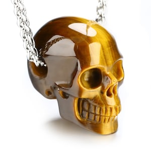 Puede incluir: Un colgante de calavera de ojo de tigre dorado en una cadena de plata. La calavera está tallada con detalles intrincados, incluyendo cuencas de los ojos, una nariz y una boca.
