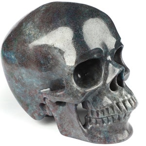 May include: A detailed, side-profile view of a decorative skull. The skull is a mix of gray, blue, and maroon hues, with a polished, textured surface. The eye sockets, nasal cavity, and teeth are clearly defined, showcasing the intricate craftsmanship.