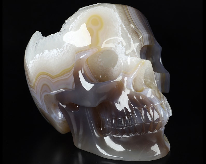 May include: A white, grey, and brown agate skull sculpture. The skull is carved from a single piece of agate and has a detailed, realistic design. The agate has a natural, banded pattern.