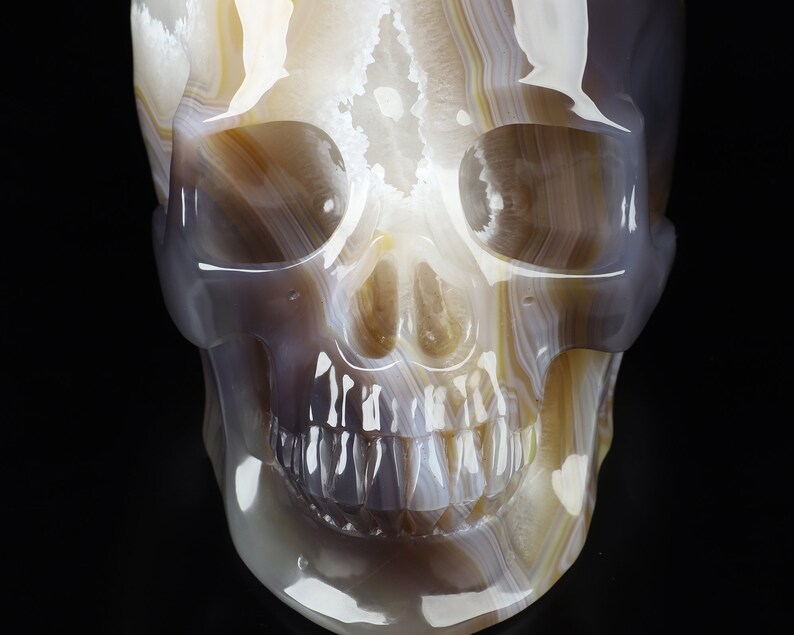 May include: A white, grey, and brown agate skull sculpture. The skull is carved with detailed features, including eye sockets, a nose, and teeth.