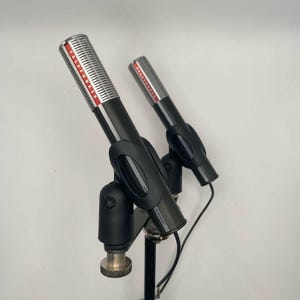 May include: Two vintage microphones with black bodies and silver mesh heads. The microphones are mounted on black stands. The brand name TELEFUNKEN is visible in red lettering on the microphone heads. The stands are labeled "Germany".