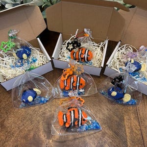 May include: Several handmade crochet fish, including orange and white clownfish and blue fish, are packaged in clear bags with blue accents. Some are in small cardboard boxes with decorative filler, suggesting a gift or craft item.