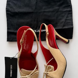 Puede incluir: Un par de zapatos de tacón destalonados en color beige con acabado texturizado, punta afilada y lazos decorativos. El forro interior y las suelas son rojos. Los zapatos cuentan con correas ajustables y vienen con una bolsa protectora negra y una etiqueta de Dolce & Gabbana.