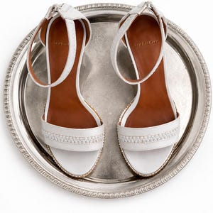 May include: A pair of white leather sandals with ankle straps, set on a silver tray. The sandals have a decorative band across the toes and are labeled "GIVENCHY". The soles are brown.