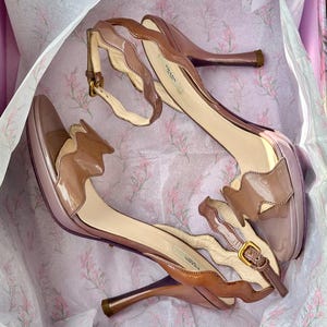 Peut inclure: Une paire de sandales à talons hauts dans les tons marron et rose, avec un design festonné sur les bords. Les sandales ont des brides de cheville avec des boucles dorées et la marque "PRADA" est visible sur la semelle intérieure.