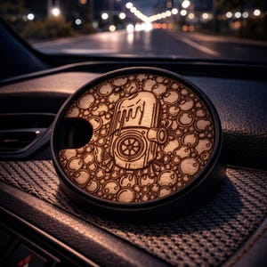 May include: A round, black car accessory with a wooden top featuring an engraved design of a mechanical component surrounded by bubbles. The accessory is resting on a textured dashboard, with a blurred city street in the background.
