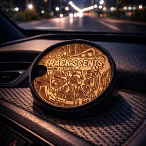 May include: A car air freshener with a wooden top featuring the text "TRACK SCENTS" and an abstract design. The air freshener is set in a black holder, resting on a car dashboard with a city night view in the background.