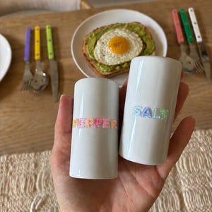 May include: Two cylindrical white ceramic shakers with flat tops. One shaker features the word PEPPER in multicolored raised letters, and the other features the word SALT in multicolored raised letters. The shakers have a smooth, glossy finish.
