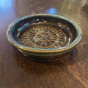 May include: A small, round ceramic dish with a dark brown interior and a raised, textured floral design. The rim is a contrasting teal-green color. The dish is likely used for serving or display.