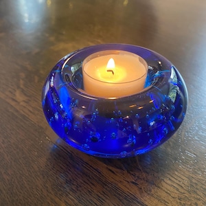 May include: A round, cobalt blue glass candle holder with a lit, cream-colored tea light candle inside. The glass has a bubbly texture and sits on a dark wooden surface. The candle flame is bright.