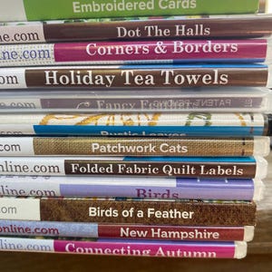 May include: A stack of clear plastic cases containing craft patterns with titles including Embroidered Cards, Dot The Halls, Corners & Borders, Holiday Tea Towels, Rustic Leaves, Patchwork Cats, Folded Fabric Quilt Labels, Birds, Birds of a Feather, New Hampshire, and Connecting Autumn.