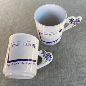 May include: Two white ceramic mugs featuring the purple text BONWIT TELLER and the phrase May I charge this to your account? Each mug has a heart-shaped handle decorated with a small cluster of purple and green floral illustrations.