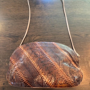 May include: A brown and black snakeskin patterned shoulder bag with a rounded top silhouette and a thin, matching shoulder strap. The bag features a textured, scale-like surface with diagonal panels of varying dark and light brown tones.