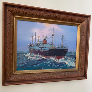 May include: Oil painting of a ship at sea, framed in a dark wood frame with ornate carvings. The ship is black and blue with red accents, sailing on turquoise waves under a blue sky with clouds and birds.