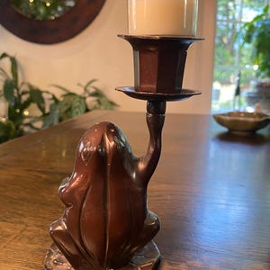 May include: A bronze-colored candle holder featuring a frog base supporting a candle cup. The candle holder has a detailed frog design with a lily pad base. The candle is in a clear glass holder.