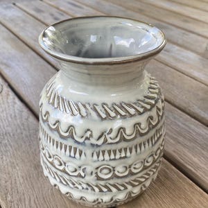 May include: A ceramic vase with a wide, flared rim and a rounded body. The exterior features a glossy, off-white glaze over a dark brown clay base, with intricate, hand-carved horizontal bands of repeating geometric patterns, including waves and circles.