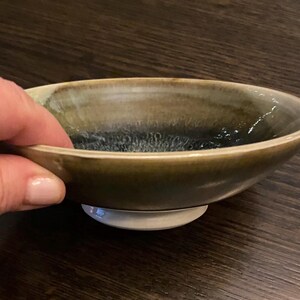May include: A small, handcrafted ceramic bowl with a unique glaze. The bowl has a dark olive green exterior that fades into a deep blue-black interior. The bowl sits on a small, white base.
