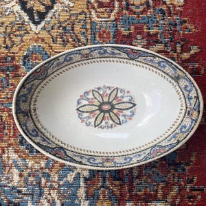 May include: An oval ceramic bowl featuring a cream-colored base with a central floral motif in black, blue, and red. The rim is decorated with a repeating mosaic-style pattern in blue, red, and tan, characterized by a visible crackle glaze finish.