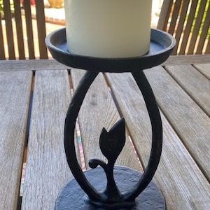 May include: A black cast iron candle holder with a circular base and a top plate. The central support features an oval frame with a decorative leaf-shaped cutout in the middle. A thick, cylindrical white pillar candle sits on the top plate.