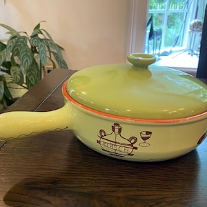 May include: A light green ceramic fondue pot with a matching lid and a single handle. The exterior features a brown graphic illustration of a bottle and a glass with the text KIRSCH printed below the bottle. The rim has an orange accent.